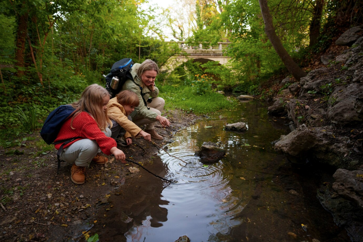 En mor sammen med to børn sidder på hug foran et vandløb