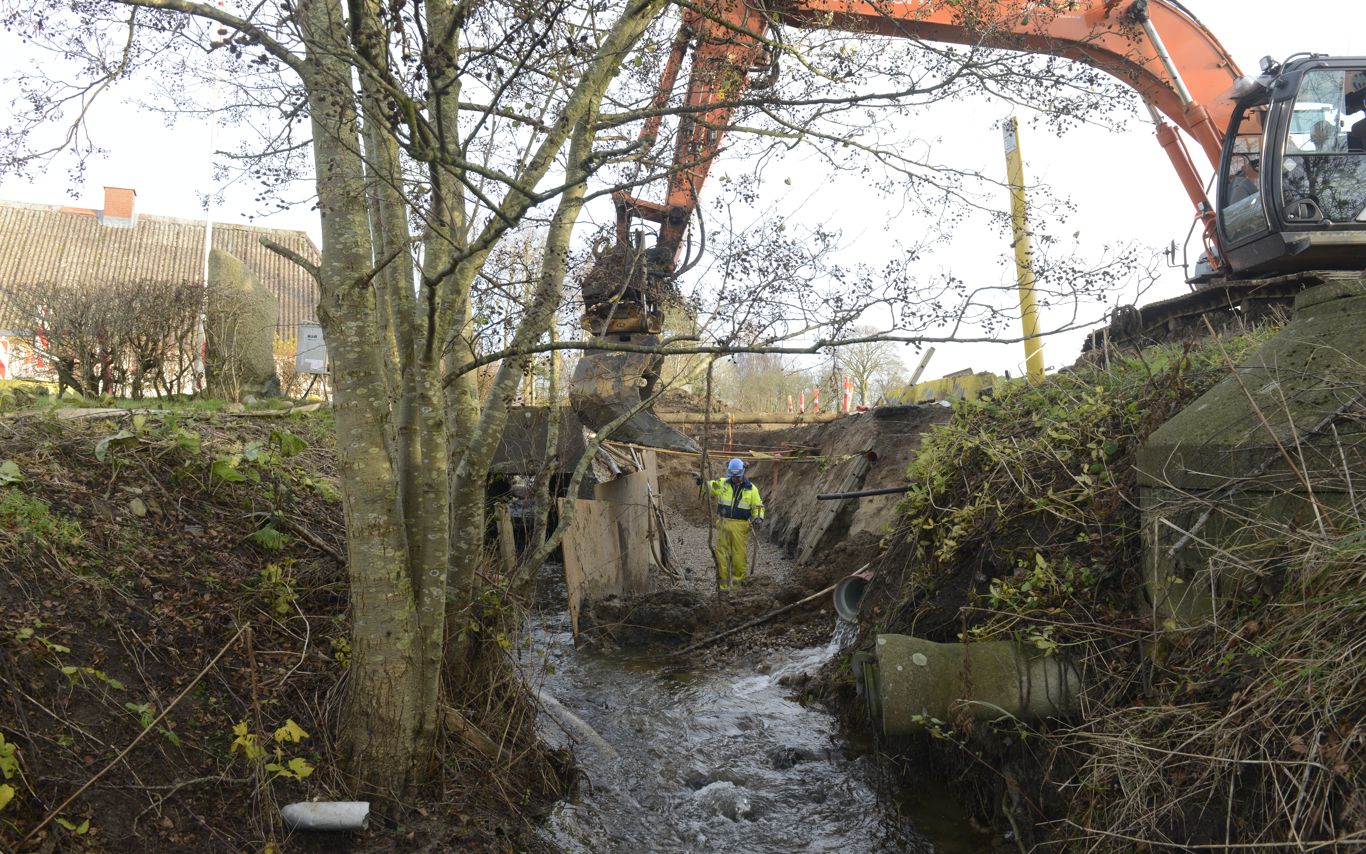 En gravko er ved at fylde sten i et vandløb (Spangebækken). En mand arbejder i vandløbet, som har stejle sider, som er afskærmet med brædder under arbejdet