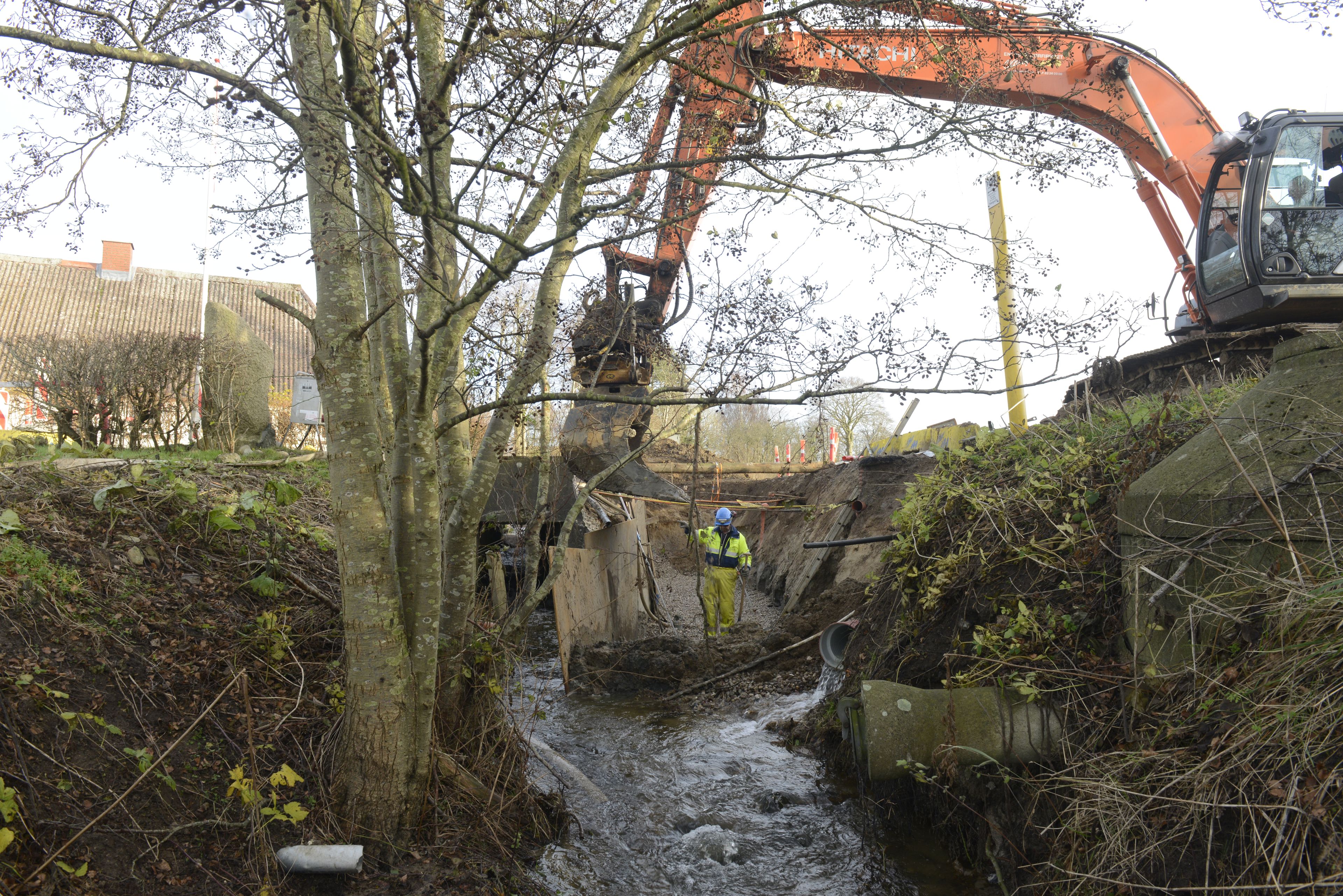 En gravko er ved at fylde sten i et vandløb (Spangebækken). En mand arbejder i vandløbet, som har stejle sider, som er afskærmet med brædder under arbejdet