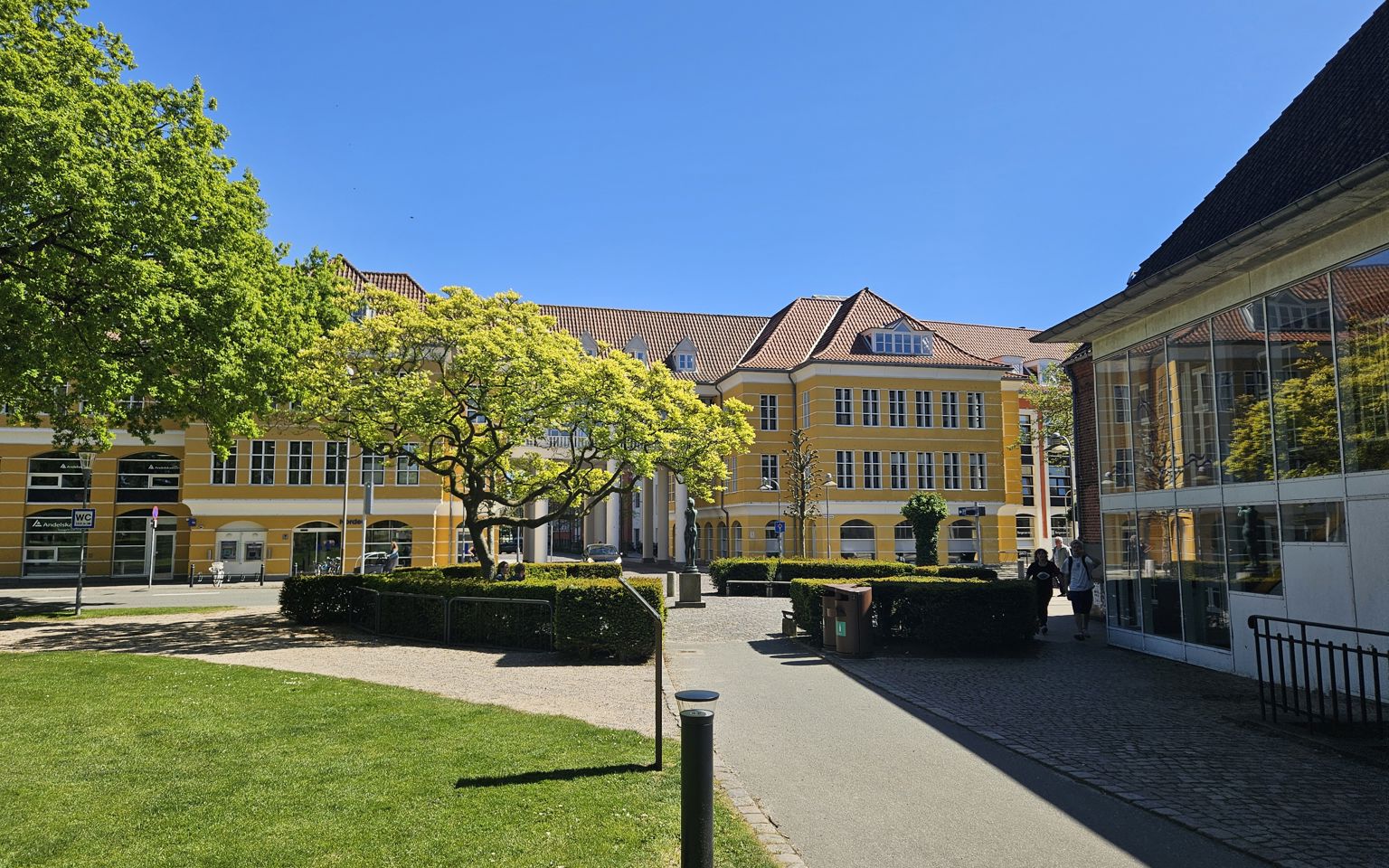 En sti med græs omkring, fører ned til en stor gul bygning med hvide vinduer. Foran bygningen er en plads omkranset af en rund hæk og et stort træ.