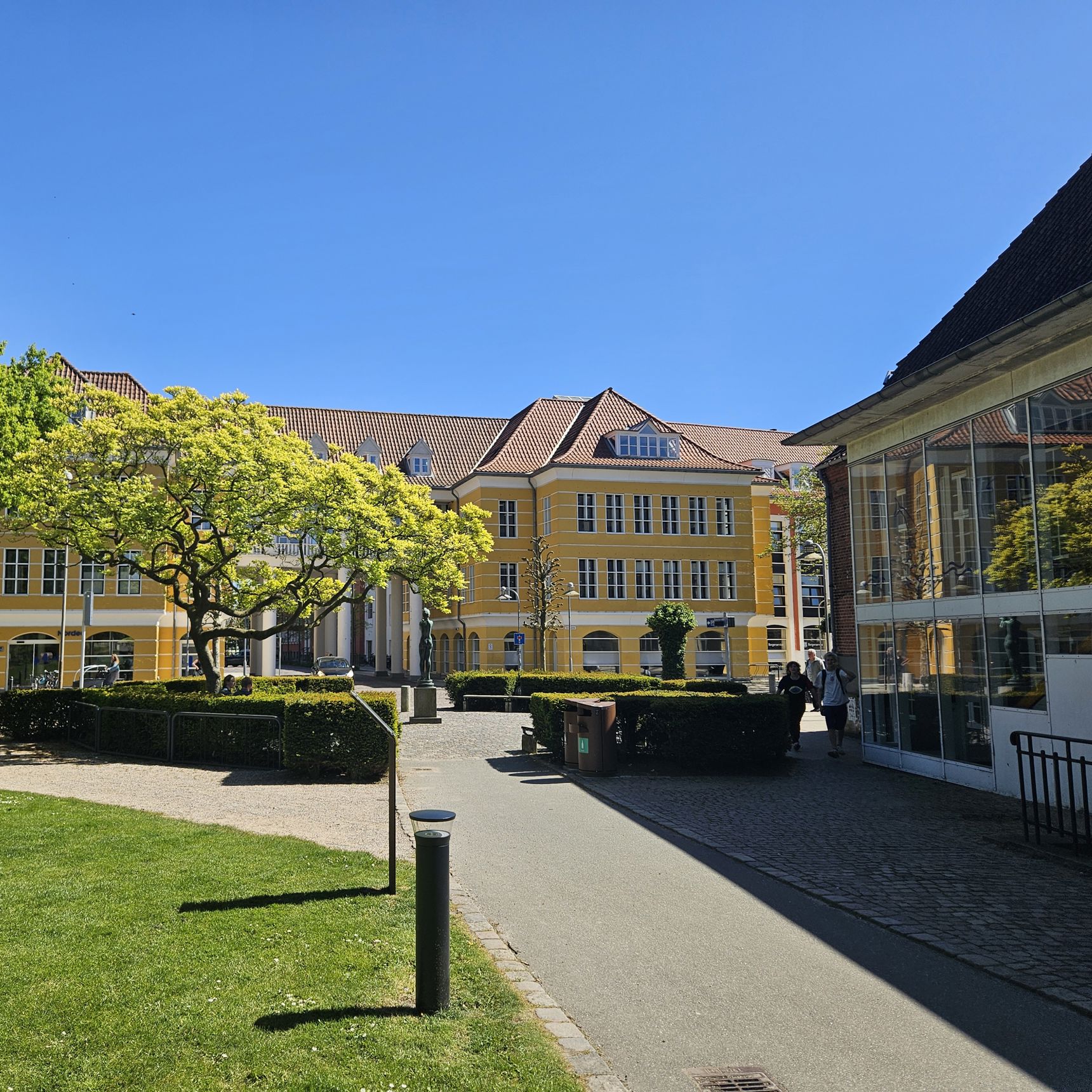 En sti med græs omkring, fører ned til en stor gul bygning med hvide vinduer. Foran bygningen er en plads omkranset af en rund hæk og et stort træ.