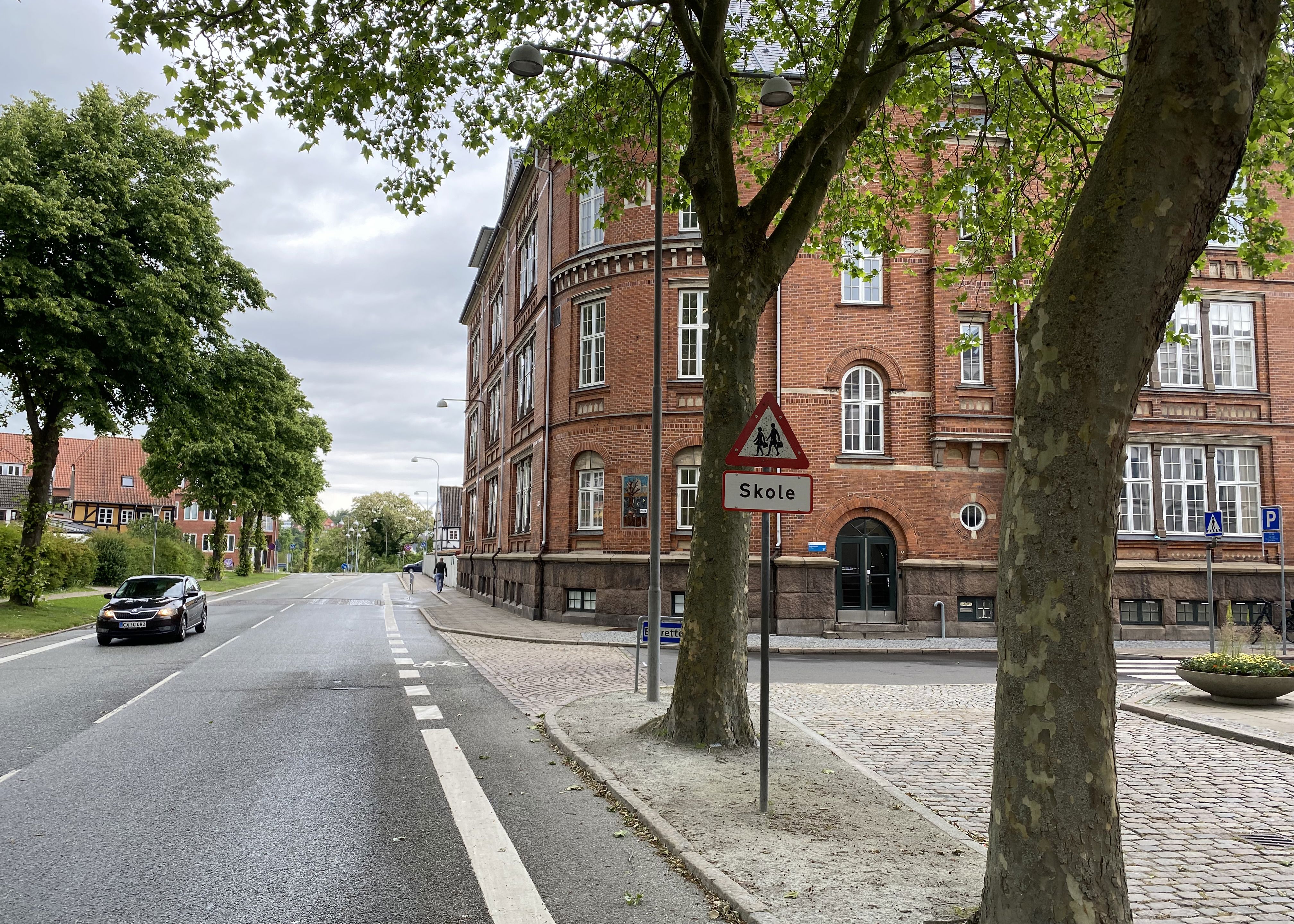 Ørkildskolen afdeling Byen - en gammel rød murstensbygning set fra vejen - udsynet er delvis blokeret med træer i forgrunden og det klassiske trekantede skilt for skole