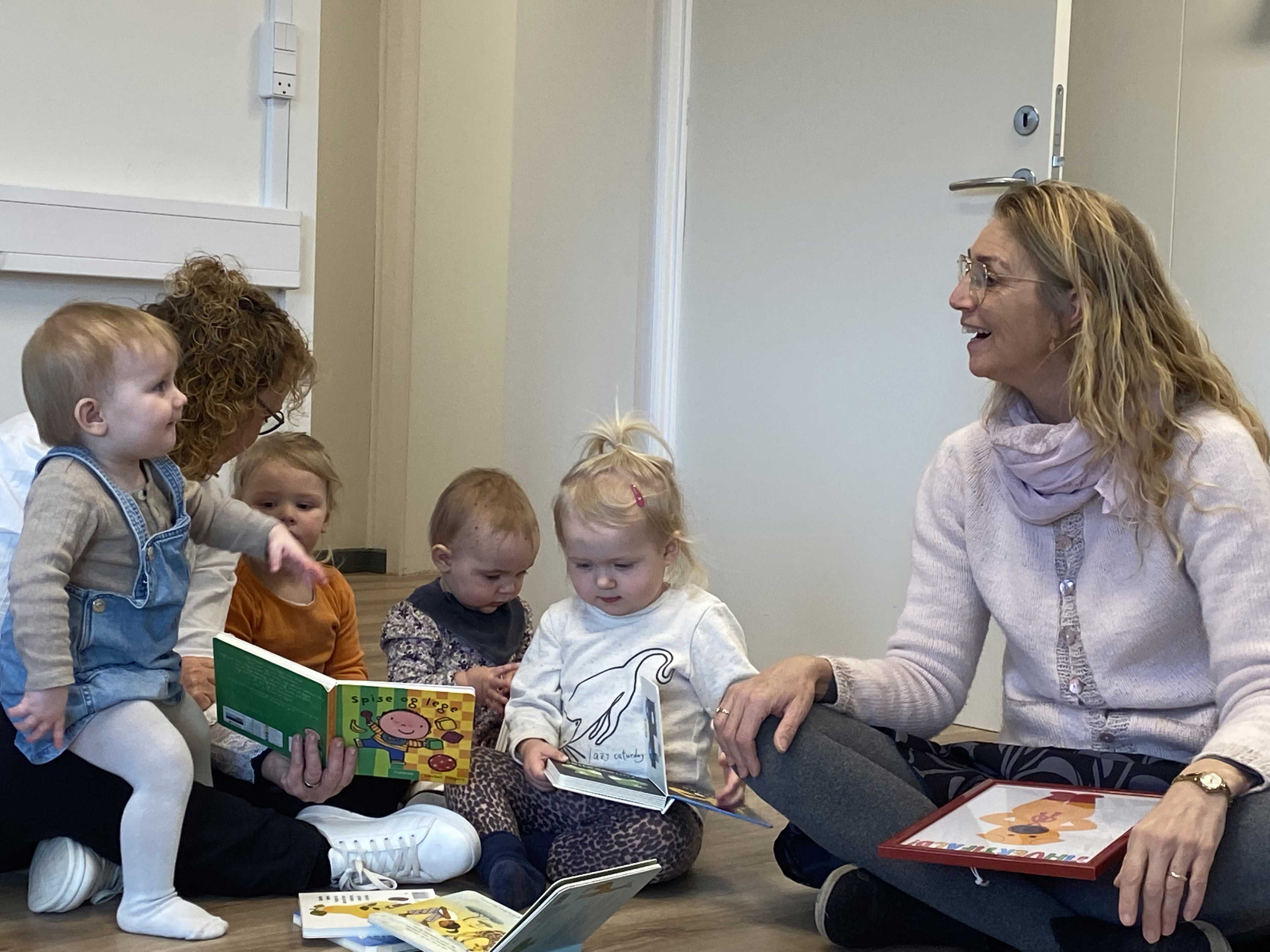 Fire små børn sidder på gulvet og kigger i bøger med to kvinder.