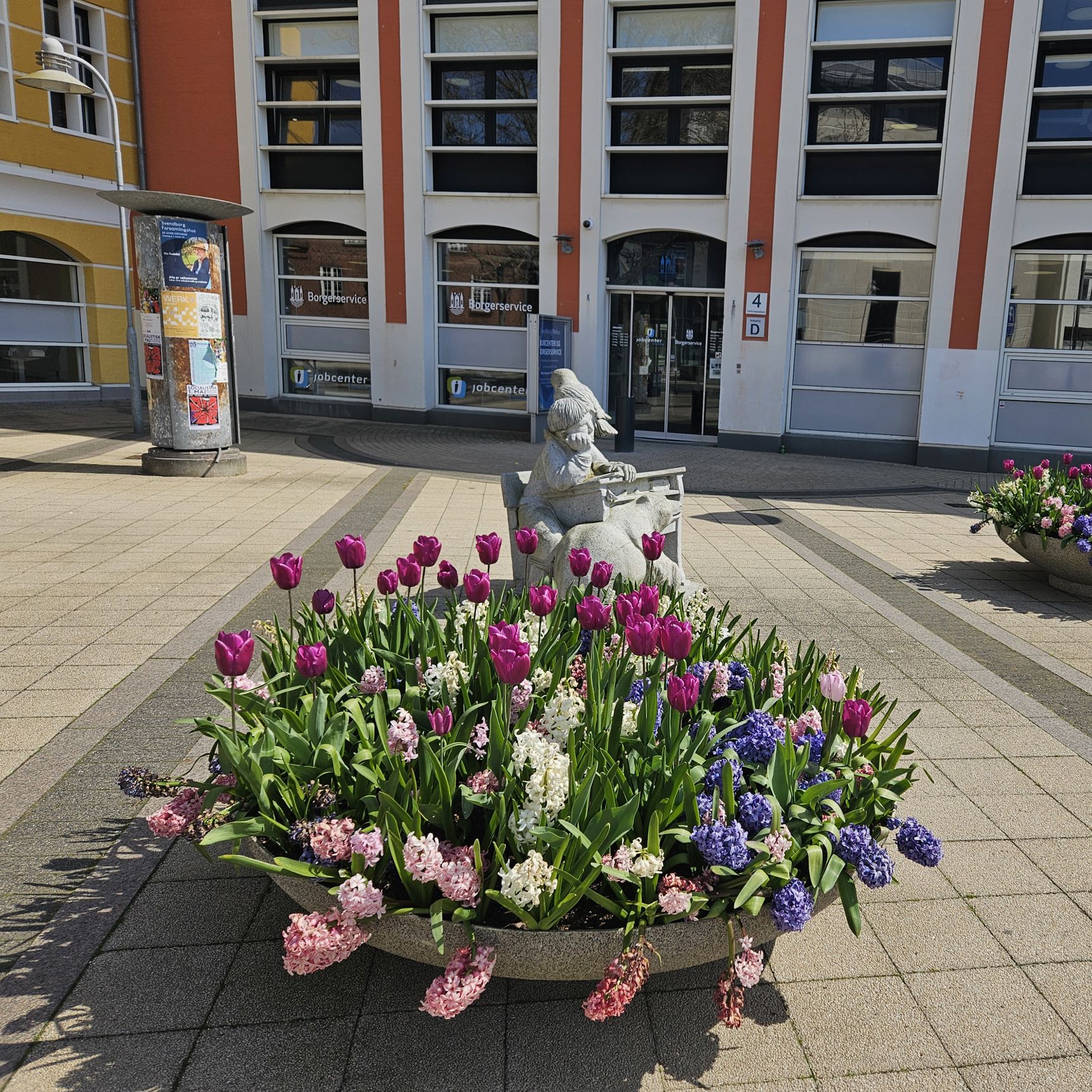 En kumme med forårsblomster foran indgangen til en stor bygning, der er hvid og rød.